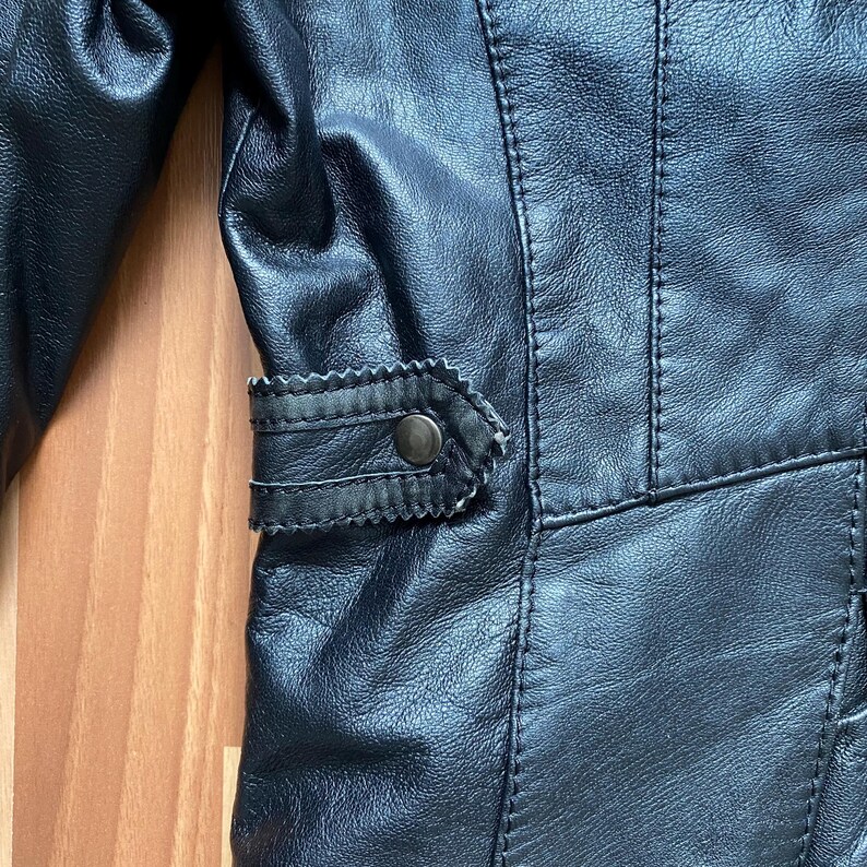K&ouml;nnte beinhalten: Nahaufnahme einer schwarzen Lederjacke. Die Jacke hat detaillierte N&auml;hte, eine dekorative Klappe mit einem Knopf und eine strukturierte Oberfl&auml;che. Das Design deutet auf einen klassischen oder Vintage-Stil hin.