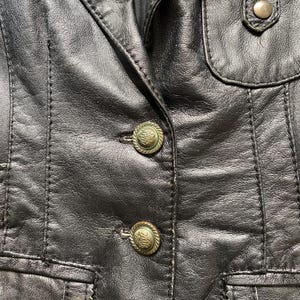 K&ouml;nnte beinhalten: Nahaufnahme einer schwarzen Lederjacke mit dekorativen N&auml;hten und zwei verzierten, goldfarbenen Kn&ouml;pfen. Die Jacke hat ein klassisches Design mit taillierter Passform und einer kleinen Tasche.