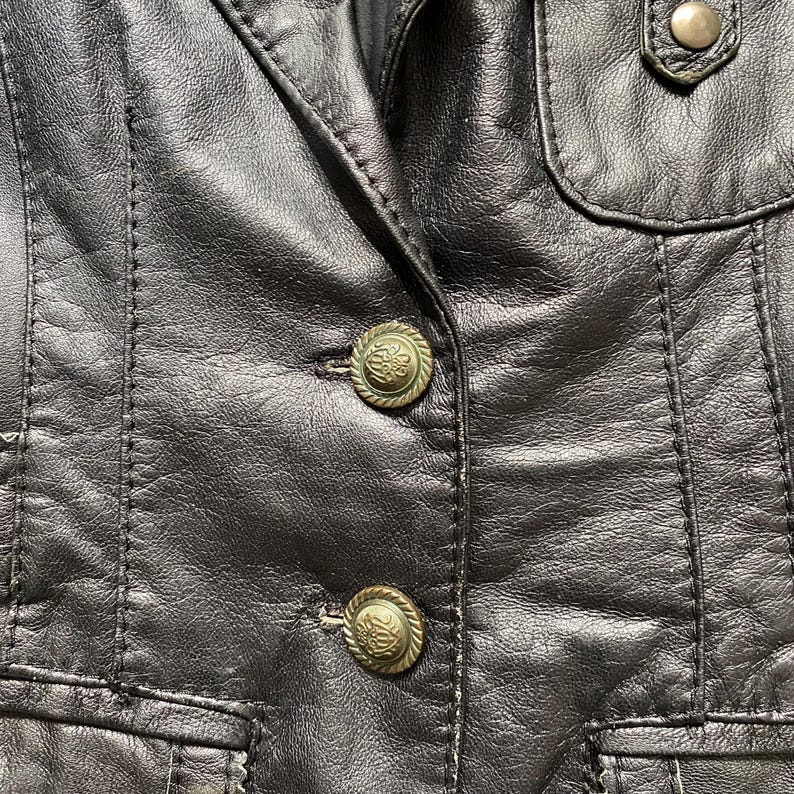 K&ouml;nnte beinhalten: Nahaufnahme einer schwarzen Lederjacke mit dekorativen N&auml;hten und zwei verzierten, goldfarbenen Kn&ouml;pfen. Die Jacke hat ein klassisches Design mit taillierter Passform und einer kleinen Tasche.