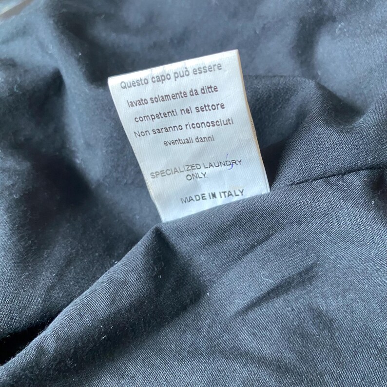 K&ouml;nnte beinhalten: Ein wei&szlig;es Etikett mit italienischem Text, darunter "SPECIALIZED LAUNDRY ONLY" und "MADE IN ITALY." Das Etikett ist an einem schwarzen Stoff befestigt. Der Text empfiehlt eine professionelle Reinigung.