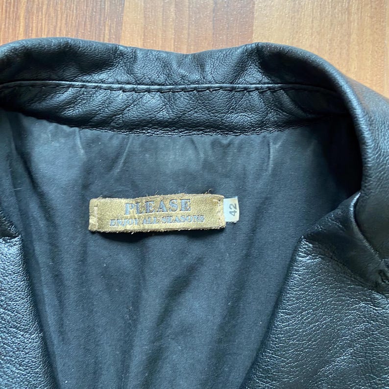 K&ouml;nnte beinhalten: Nahaufnahme eines schwarzen Lederjackenkragens. Die Jacke hat ein gestepptes Design und ein Etikett mit der Aufschrift "PLEASE" und "ENJOY ALL SEASONS". Das Etikett hat auch die Nummer "42".