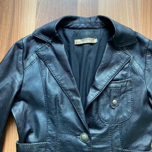 K&ouml;nnte beinhalten: Schwarzer Lederblazer mit Reverskragen, Ein-Knopf-Verschluss und kleiner Brusttasche. Der Blazer hat eine taillierte Passform und sichtbare Nahtdetails. Das Etikett lautet "PLEASE".