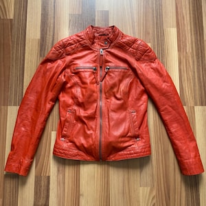 May include: A vibrant red leather jacket with a stand-up collar and a full-length zipper. The jacket features quilted shoulder detailing, two zippered chest pockets, and two side pockets. The jacket is a size medium.