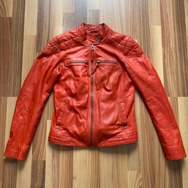 May include: A vibrant red leather jacket with a stand-up collar and a full-length zipper. The jacket features quilted shoulder detailing, two zippered chest pockets, and two side pockets. The jacket is a size medium.