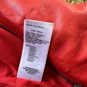 May include: Close-up of a red leather garment with a zipper. A white tag displays material information in multiple languages, including "Leder/leather" and "100% Polyester." The tag also includes the manufacturer's address in Germany.