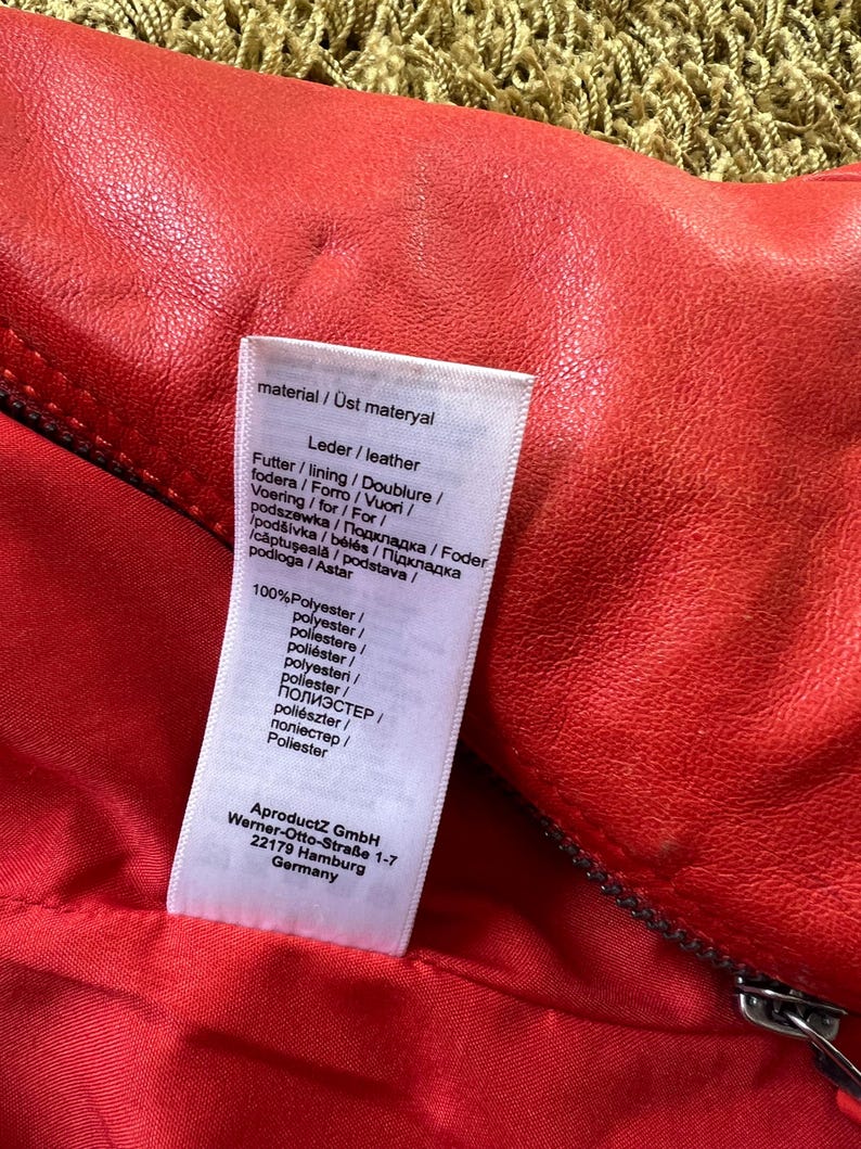 May include: Close-up of a red leather garment with a zipper. A white tag displays material information in multiple languages, including "Leder/leather" and "100% Polyester." The tag also includes the manufacturer's address in Germany.