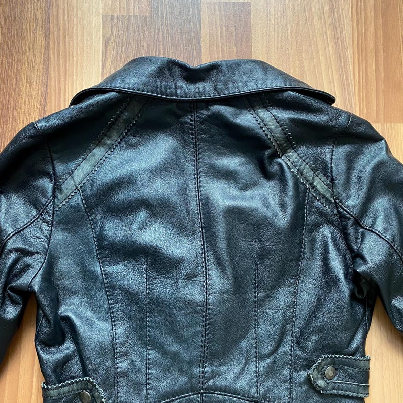 K&ouml;nnte beinhalten: Schwarze Lederjacke mit Reverskragen und dekorativen N&auml;hten. Die Jacke hat eine taillierte Silhouette mit einem taillierten R&uuml;cken und Kn&ouml;pfen in der N&auml;he der Taille. Das Leder scheint leicht abgenutzt zu sein, was ihm einen Vintage-Look verleiht.