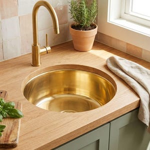 Pode incluir: Uma pia de cozinha com uma torneira dourada e uma bacia de pia redonda dourada embutida em uma bancada de madeira. Um pequeno vaso de terracota com uma planta de alecrim está sobre a bancada, junto com uma tábua de corte de madeira com manjericão.