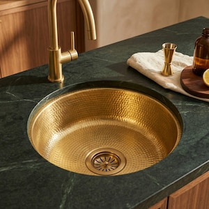 May include: A hammered gold kitchen sink with a matching faucet. The sink is round and set into a dark green countertop. A small wooden cutting board with a lemon and a brown bottle are visible.