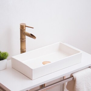 May include: A modern bathroom sink with a rectangular white basin and a bamboo-style brass faucet. A small potted plant and a towel rack with a white towel are also visible. The sink is on a white countertop.