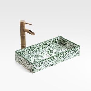 May include: A rectangular ceramic sink with a green and white floral pattern, paired with a bamboo-style brass faucet. The sink has a central drain and is set against a white background. The faucet is positioned to the left of the sink.