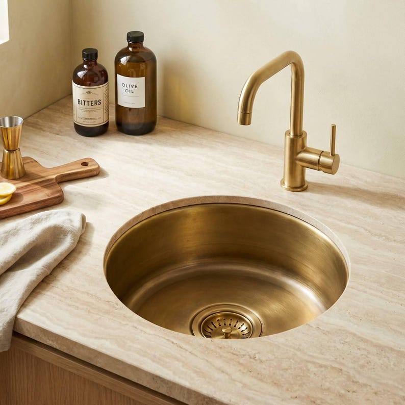 TerraFez handcrafted brass round sink installed in light travertine countertop with brushed gold faucet, cocktail bitters, olive oil bottles, cutting board, and lemon slices in a styled home bar scene.
