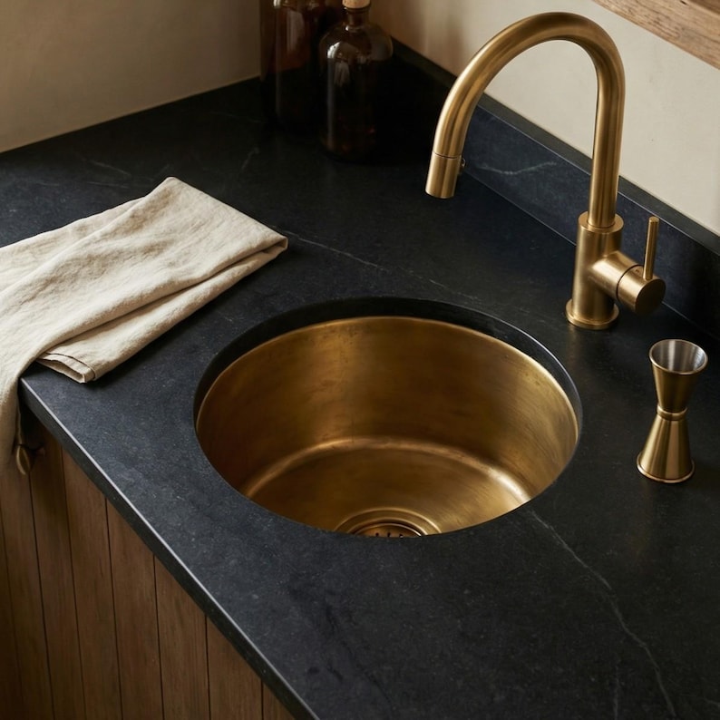 Handcrafted solid brass round bar sink by TerraFez installed in dark soapstone countertop with brushed gold faucet, linen towel, and wooden cabinetry in a modern home bar setting.