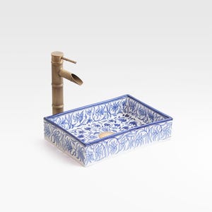Könnte beinhalten: Ein rechteckiges Keramikwaschbecken mit blau-weißem Blumenmuster und einem Wasserhahn aus Messing im Bambus-Stil. Das Waschbecken hat einen goldfarbenen Abfluss und steht vor einem weißen Hintergrund.