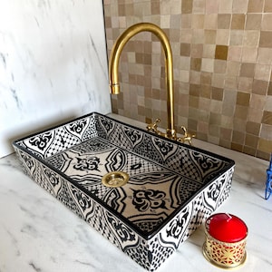 Könnte beinhalten: Ein weißes Keramikwaschbecken mit einem geometrischen Muster in Schwarz und Weiß. Das Waschbecken ist rechteckig und hat einen goldenen Wasserhahn.