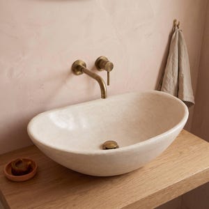 May include: An oval-shaped off-white stone vessel sink with a smooth finish and a central drain. A wall-mounted brass faucet with a single lever handle is positioned above the basin. A small round dish with a brown soap bar sits nearby.