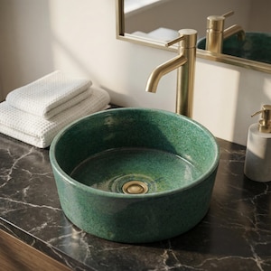 May include: A green ceramic vessel sink with a gold-coloured drain and tap. The sink is round and sits on a dark countertop. White towels are stacked to the left, and a mirror is in the background.
