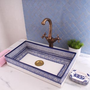 Op de afbeelding: Een rechthoekige badkamer wastafel van wit keramiek met een blauw en wit geometrisch patroon rond de rand. De wastafel heeft een messing kraan met twee handvatten en een afvoer in het midden. Een kleine witte keramische zeepbak met een blauw en wit geometrisch patroon bevindt zich in de rechter benedenhoek van de afbeelding.