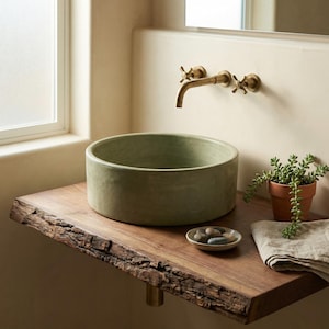 Può includere: Un lavabo rotondo verde salvia è appoggiato su una mensola in legno naturale con bordo vivo. Un piattino con pietre, una pianta in vaso e un asciugamano di lino piegato sono anch'essi sulla mensola. Il rubinetto è color ottone.