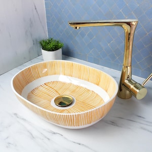 Puede incluir: Un lavabo ovalado de cerámica blanca con un patrón de rayas doradas. El lavabo tiene una tapa de drenaje dorada y está colocado sobre una encimera de mármol blanco.