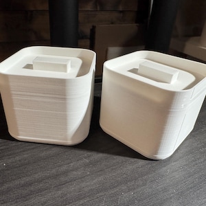 May include: Two white, square containers with lids. Each container has a rectangular block on top of the lid. The containers are made of a ridged material and sit on a dark surface.