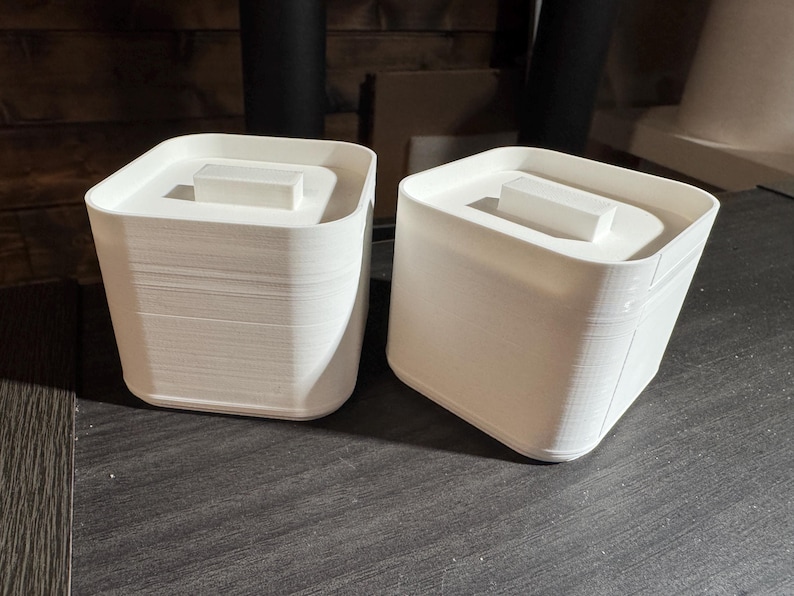 Puede incluir: Dos recipientes blancos cuadrados con tapas. Cada recipiente tiene un bloque rectangular en la parte superior de la tapa. Los recipientes est&aacute;n hechos de un material estriado y se asientan sobre una superficie oscura.