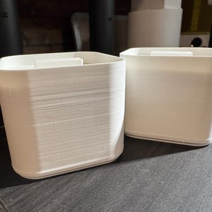 Puede incluir: Dos recipientes blancos y rectangulares con un aspecto texturizado y en capas. Cada recipiente tiene una peque&ntilde;a pieza rectangular en la parte superior. Los recipientes est&aacute;n sobre una superficie oscura.