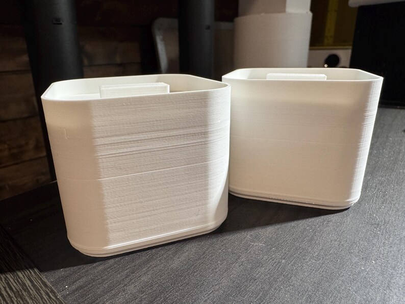 Puede incluir: Dos recipientes blancos y rectangulares con un aspecto texturizado y en capas. Cada recipiente tiene una peque&ntilde;a pieza rectangular en la parte superior. Los recipientes est&aacute;n sobre una superficie oscura.