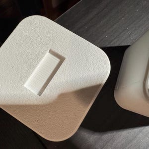 Puede incluir: Dos objetos cuadrados de color blanquecino con esquinas redondeadas. Un objeto tiene una hendidura rectangular. Los objetos tienen una superficie texturizada. Los objetos son probablemente para exhibici&oacute;n o almacenamiento.