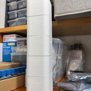 Puede incluir: Un objeto rectangular blanco y alto, con esquinas redondeadas, probablemente impreso en 3D, est&aacute; apilado en una estanter&iacute;a de madera. El objeto est&aacute; compuesto por varias secciones apiladas id&eacute;nticas. El fondo incluye contenedores de almacenamiento y componentes electr&oacute;nicos.