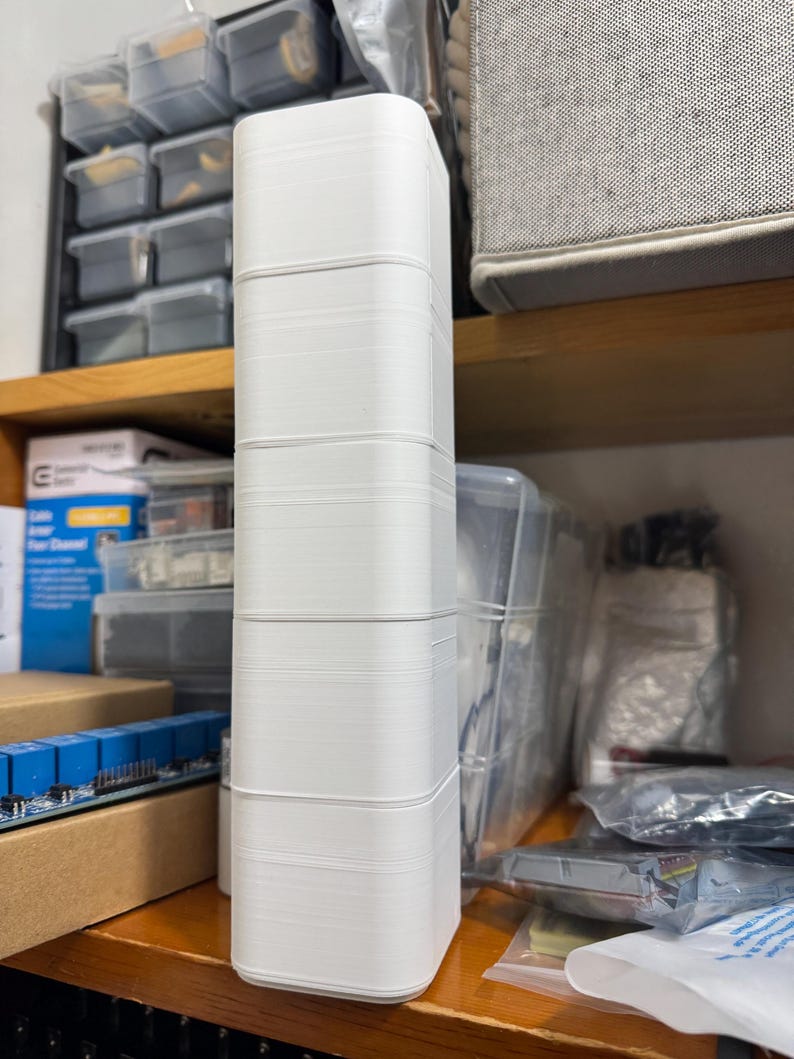 Puede incluir: Un objeto rectangular blanco y alto, con esquinas redondeadas, probablemente impreso en 3D, est&aacute; apilado en una estanter&iacute;a de madera. El objeto est&aacute; compuesto por varias secciones apiladas id&eacute;nticas. El fondo incluye contenedores de almacenamiento y componentes electr&oacute;nicos.