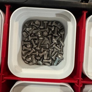 Puede incluir: Primer plano de un recipiente cuadrado blanco lleno de numerosos tornillos plateados peque&ntilde;os. Los tornillos tienen cabeza plana y est&aacute;n bien ordenados. El recipiente forma parte de un sistema de almacenamiento rojo y blanco.