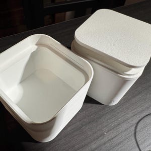 Puede incluir: Dos recipientes blancos de forma cuadrada con esquinas redondeadas y tapas texturizadas. Un recipiente est&aacute; abierto, revelando su interior vac&iacute;o. El otro recipiente est&aacute; cerrado, con la tapa ligeramente entreabierta. Los recipientes est&aacute;n sobre una superficie oscura.