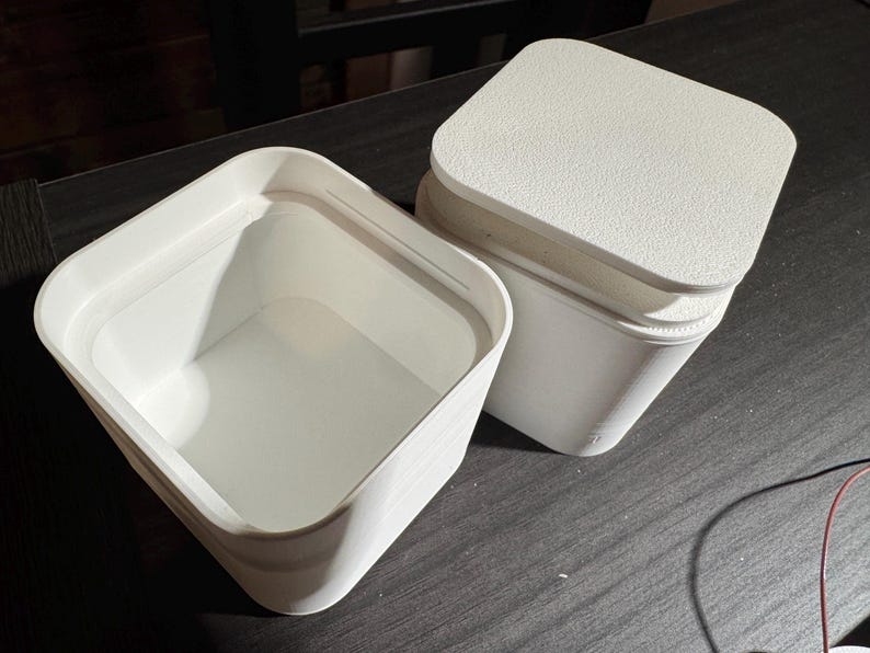 Puede incluir: Dos recipientes blancos de forma cuadrada con esquinas redondeadas y tapas texturizadas. Un recipiente est&aacute; abierto, revelando su interior vac&iacute;o. El otro recipiente est&aacute; cerrado, con la tapa ligeramente entreabierta. Los recipientes est&aacute;n sobre una superficie oscura.