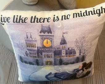 May include: Decorative pillow with a castle illustration and the text "Live like there is no midnight" in black. The castle has a large clock face and is set against a purple and blue background. The pillow is white.