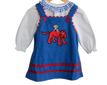 May include: A blue corduroy toddler dress with red trim and a white long-sleeved shirt. The dress features an embroidered image of a red dog with a girl riding on its back. The dress is hanging on a wooden hanger.