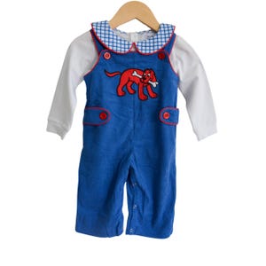 May include: Blue corduroy baby overalls with a white long-sleeve shirt. The overalls feature a red cartoon dog holding a bone, red buttons, and red trim. The collar is white with a blue and white checkered pattern.