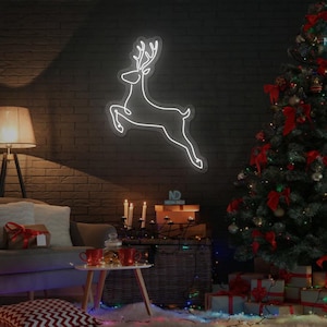 Op de afbeelding: Een wit neon rendierbord aan een donkere bakstenen muur. Rechts staat een versierde kerstboom met rode strikken en ornamenten. Een bank, cadeaus en een kleine tafel met drankjes maken de feestelijke scène compleet.
