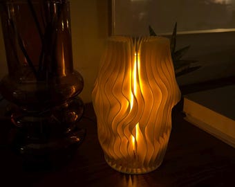 May include: A white, wooden lamp with a wavy, pleated design. The lamp is lit and casting a warm glow.