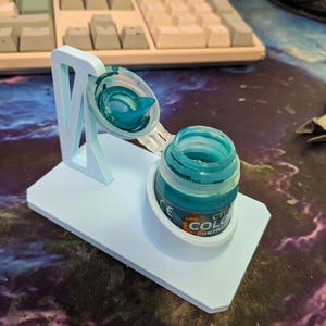 Puede incluir: Un soporte para bote de pintura de color azul claro impreso en 3D con un brazo de soporte vertical y una base con corte circular. El soporte contiene un bote de pintura verde azulado abierto con tapa abatible y una etiqueta parcial.