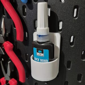 Puede incluir: Un soporte de plástico blanco con una botella de pegamento Bison Lock Bond dentro. La botella es negra con una etiqueta azul y una tapa blanca.