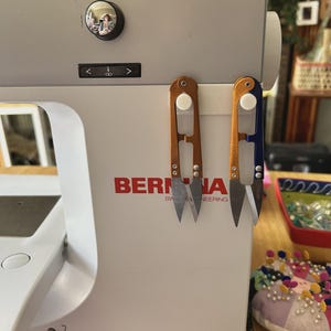 Puede incluir: Dos cortahilos con mangos de color naranja y azul metálico están sujetos al lateral de una máquina de coser Bernina blanca. Los cortahilos cuentan con hojas de metal afiladas y puntiagudas, montadas sobre una banda magnética blanca.