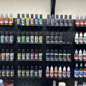 Peut inclure: Une collection de flacons de peinture acrylique de différentes couleurs, soigneusement organisés sur des étagères noires. Les flacons sont exposés sur un panneau perforé noir, présentant une variété de nuances pour les projets artistiques.