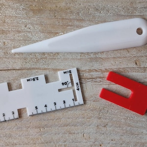 Op de afbeelding: Een witte plastic vouwbeen, een witte plastic naaimaat met markeringen van 1 cm tot 4 cm, en een klein rood U-vormig geleidingsgereedschap van plastic. De witte maat heeft gedrukte afmetingen en uitsparingen voor nauwkeurige uitlijning.