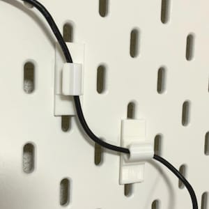 以下が含まれることがあります： 白いペグボードの壁に付けられた、白いプラスチック製のケーブルクリップ2つ。黒い電気コードを固定しています。