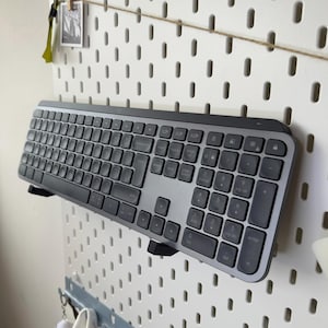 Könnte beinhalten: Eine schwarze und silberne drahtlose Tastatur mit einem Ziffernblock und Funktionstasten. Die Tastatur ist an einer weißen Lochwand montiert.