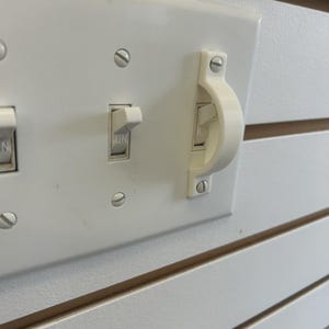 May include: A white plastic light switch guard mounted over a toggle switch on a white wall plate. The guard is a semi-circular bracket secured with two screws, designed to prevent accidental operation of the switch which is currently in the ON position.