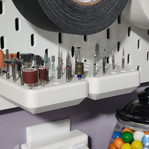 Op de afbeelding: Een wit pegboard met twee witte planken met diverse boorbits. De boorbits hebben verschillende vormen en kleuren, waaronder oranje, bruin en zilver. Rollen tape bevinden zich boven de planken.