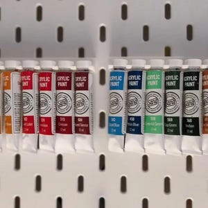 May include: A collection of acrylic paint tubes in various colors, including yellow, red, blue, and green, displayed on a white pegboard. Each tube is labeled with a color name and number, and the brand name "Sang".