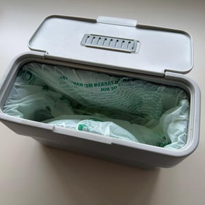Puede incluir: Un cubo de basura de plástico gris con tapa y un forro de plástico blanco. El forro tiene texto verde que dice "DE ROT AFVALKEN ME HANTEREN"